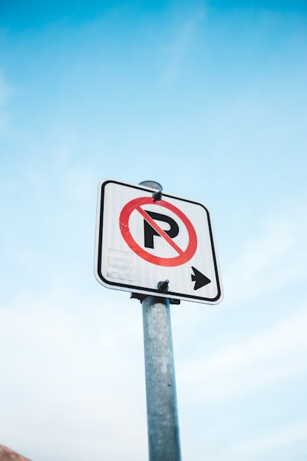 A round traffic sign mounted on a black metal bracket attached to a red brick wall, indicating parking restrictions in the area. The sign features a red border with a blue circle and a red diagonal li