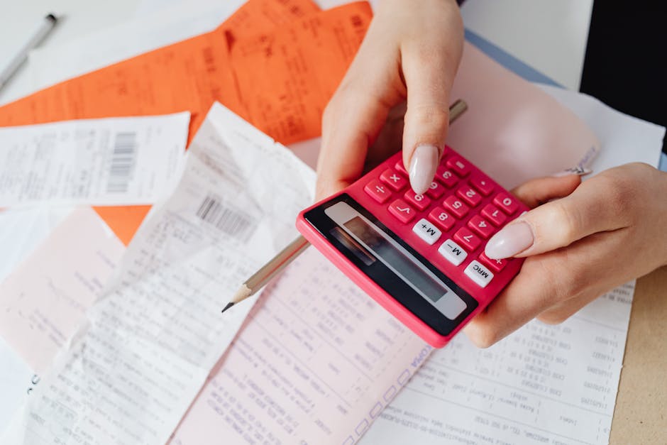 A person, wearing a light blue shirt, is seated at a desk surrounded by various receipts, invoices, and paper documents. They are using a small pink calculator, pressing buttons with their right hand,