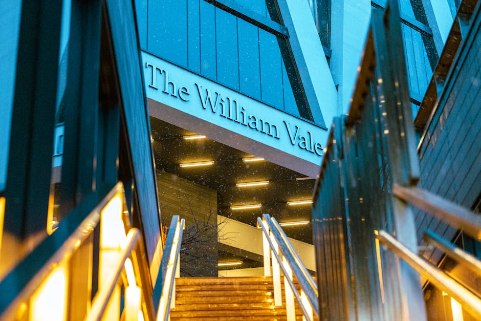 View of the exterior of The William Vale hotel at night, showing illuminated signage on the building's blue facade. The entrance features a staircase with wooden steps and white handrails, leading up from pavement to the doorway. Surrounding the staircase are metal railings and glass panels, with lighting fixtures providing ambient illumination. The image captures part of the building's modern architectural design, with exterior walls composed of blue panels and large glass windows reflecting nearby structures. The scene is focused on the entrance area, which appears prepared for a home relocation or furniture transport process by Removal Company Maida Vale, with visible elements suggesting recent packing or loading activities in a professional moving context.