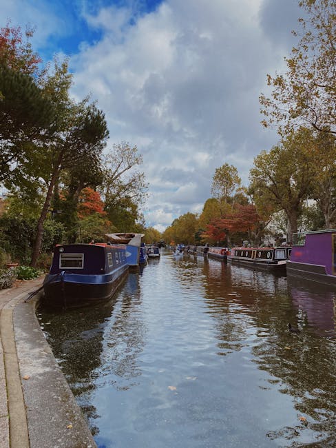 A canal scene with several narrowboats moored along the bank, featuring a mix of colours including blue, purple, and black. The water reflects the overcast sky, which has patches of blue and white clouds. Tall trees with green and orange autumn foliage line both sides of the canal, indicating a seasonal change. On the left, a paved walkway runs parallel to the water, extending into the distance, suggesting a quiet residential or leisure area. The presence of boats and the calm water imply a setting suitable for home relocation or vessel transport, while the overall scene depicts typical elements involved in house removals or moving services in a canal-side environment. The image is captured during daylight with natural lighting, emphasizing the serene atmosphere of the area. The setting highlights the importance of careful planning and transport logistics for moving household items, as represented by the peaceful yet functional environment managed by Removal Company Maida Vale.
