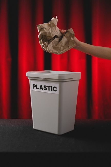 A large green wheelie bin situated outside near a dark brick wall, filled with cardboard boxes and red plastic bags containing moving supplies. The bin, which is part of the storage and disposal proce