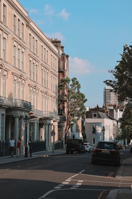 A street scene in Maida Vale featuring a traditional church with a tall clock tower and ornate architectural details, surrounded by leafy green trees and a row of parked cars along the pavement. In th