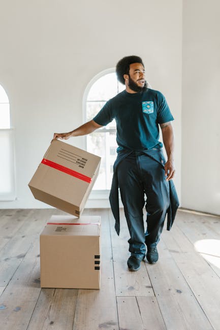 A man with dark curly hair, a beard, and dark skin is inside a bright, empty room with large arched windows, actively engaged in packing or preparing for a home relocation. He is wearing a dark blue t