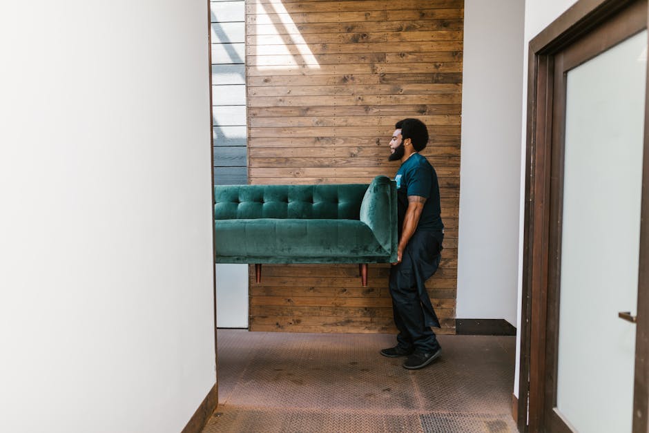 A man with dark curly hair, a beard, and dark skin is inside a bright, empty room with large arched windows, actively engaged in packing or preparing for a home relocation. He is wearing a dark blue t
